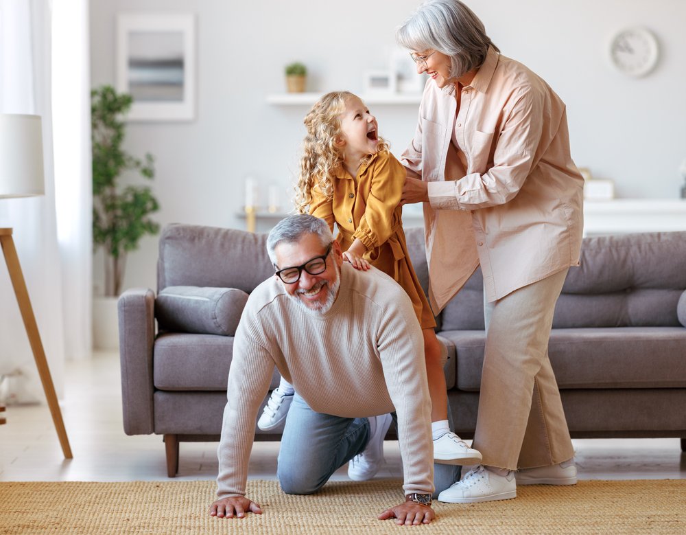 Active Senior Couple Grandparents Playing With Cute Little Girl Granddaughter In Living Room