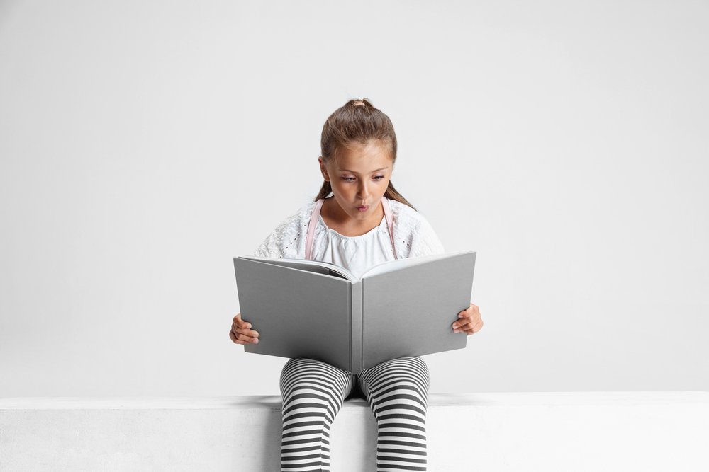 Portrait Of Cute Caucasian Girl, Teen Sitting And Reading Big Book, Magazine Isolated On Gray Studio Background.