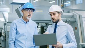 Head Of The Department Holds Laptop And Discusses Product Details With Chief Engineer. They Wear Hard Hats And Work At The Modern Factory.