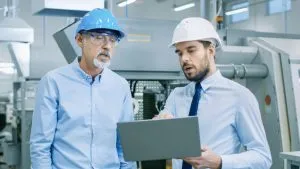 Head Of The Department Holds Laptop And Discusses Product Details With Chief Engineer. They Wear Hard Hats And Work At The Modern Factory.