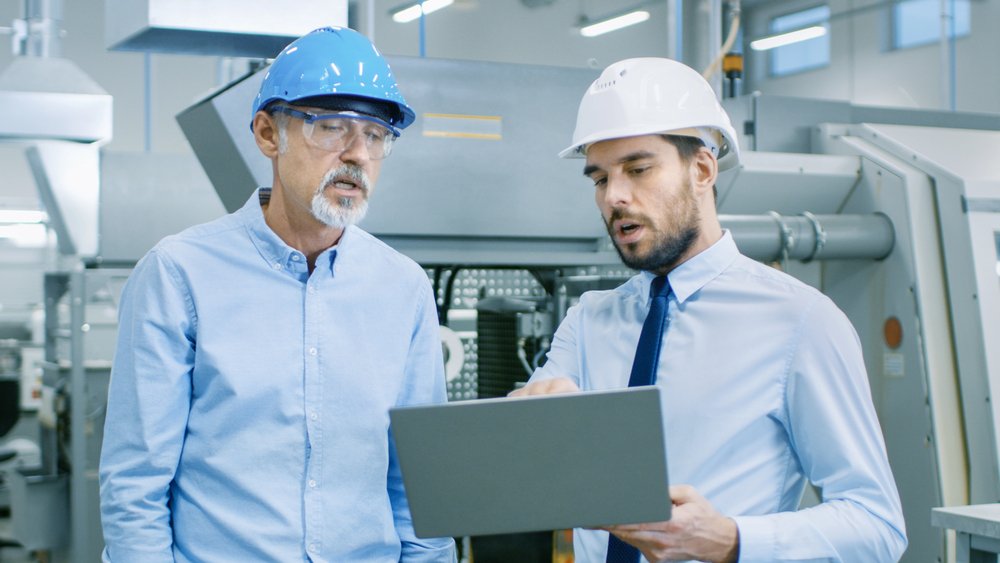 Head Of The Department Holds Laptop And Discusses Product Details With Chief Engineer. They Wear Hard Hats And Work At The Modern Factory.